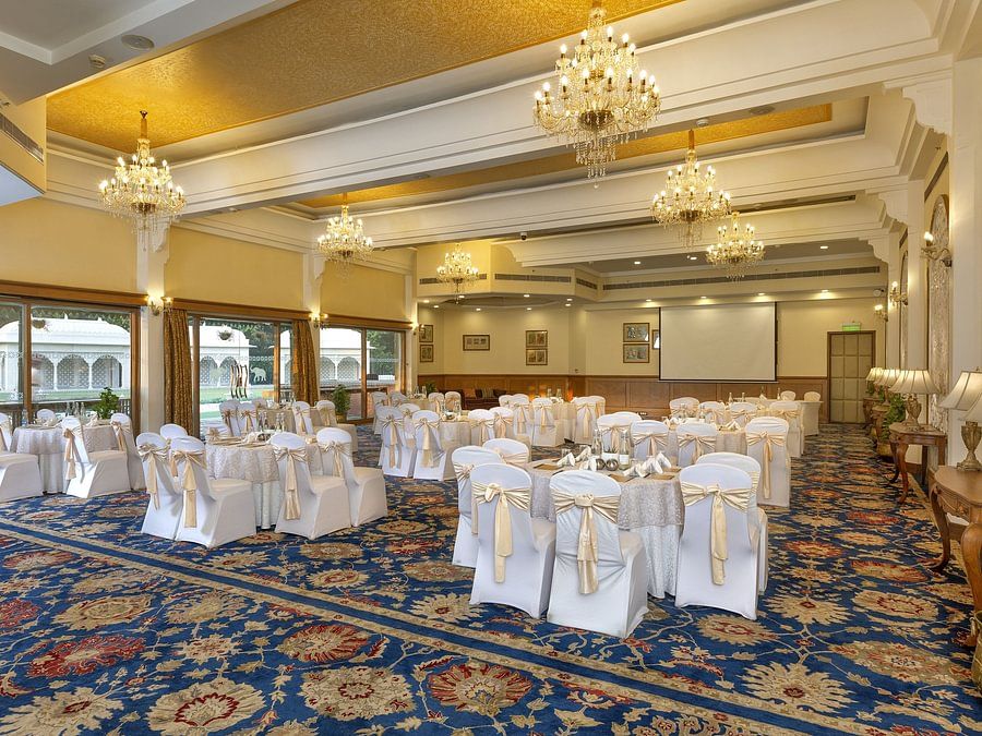 Elegant banquet hall with chandeliers, round tables draped in white, and a patterned blue carpet - Heritage Village Resorts & Spa.