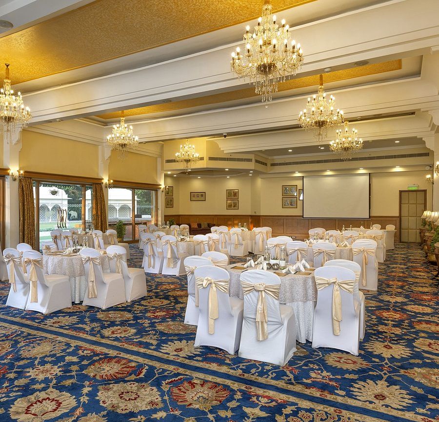 Elegant banquet hall with chandeliers, round tables draped in white, and a patterned blue carpet - Heritage Village Resorts & Spa.