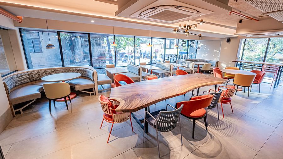 A diagonal view of a large rectangular table with chairs on both sides and a round table with chairs around it, set against glass panels in the background at Indie Stays Mumbai International Airport.