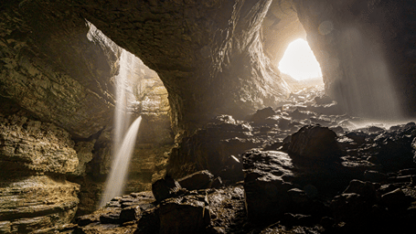 A waterfall cascades inside a vast limestone cave, where sunlight filters through natural openings onto rocky terrain and ancient formations.