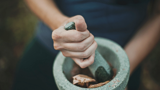 Hands hold a pestle, grinding herbs and possibly roots in a stone mortar, suggesting the preparation of traditional medicine or remedies.