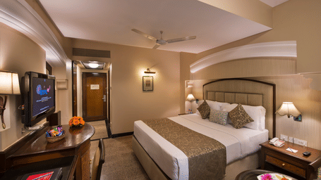 An executive room at Lords Inn Vadodara, featuring a king size bed, bedside tables and lamps, a table with snacks, a mounted T.V. and a door opening to a hallway.