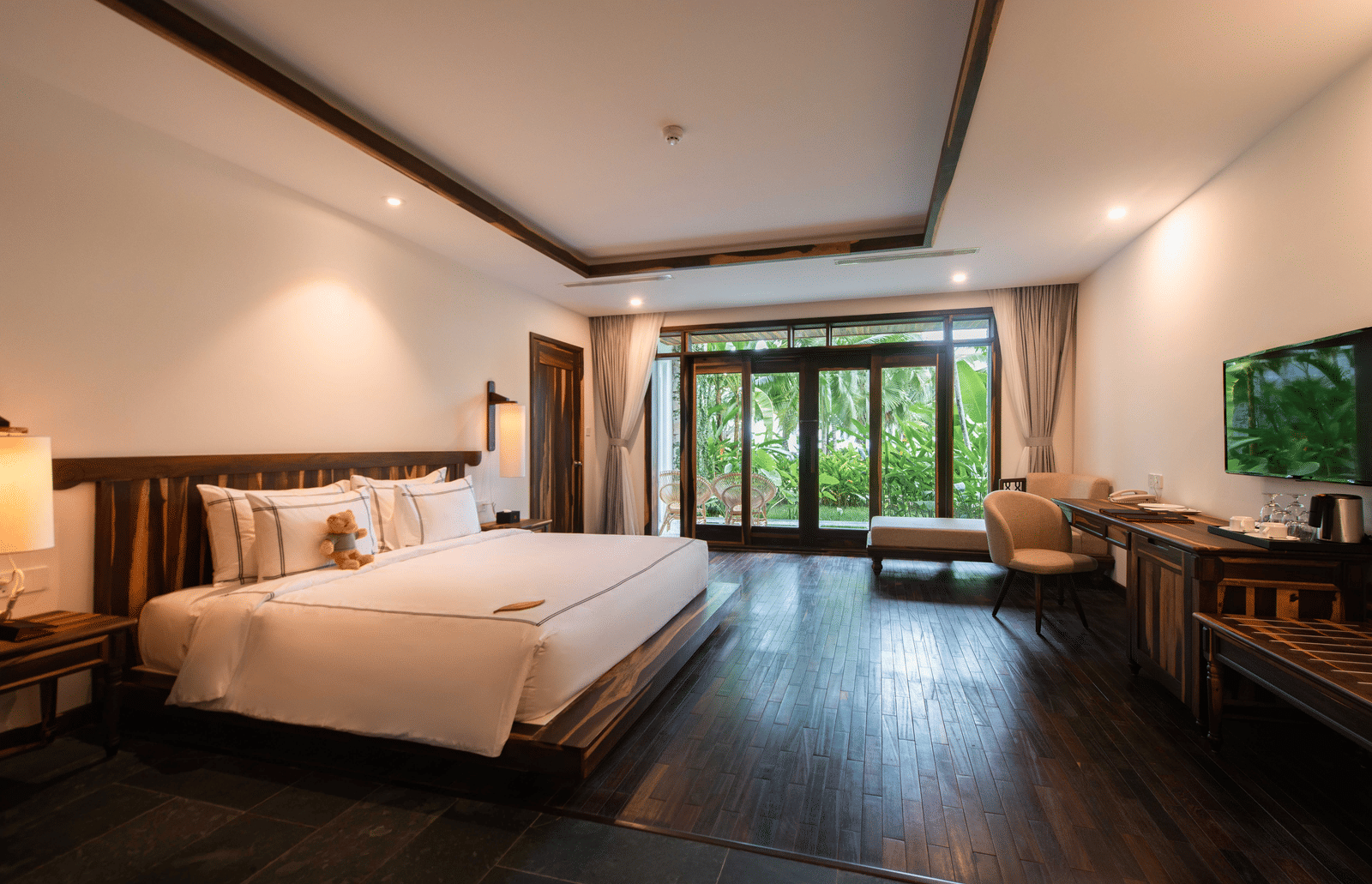 Dark floors, bedside stand, TV, reading chair, sliding glass doors, curtain and patio with garden view in king bed in Ocean View Premium at Alibu Resort Nha Trang, Vietnam.
