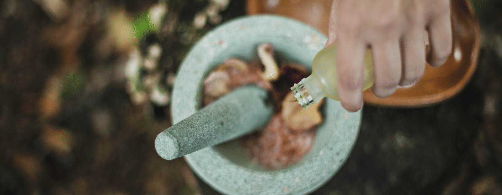 A person pours liquid into a stone mortar and pestle while preparing natural ingredients on a wooden surface.