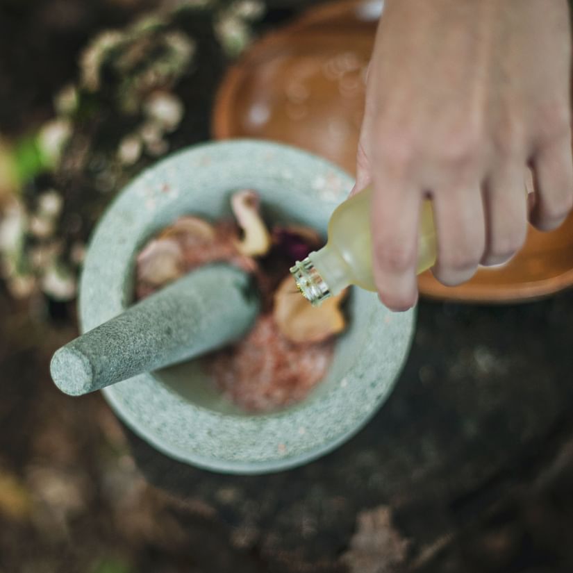 A hand pours liquid from a small bottle into a stone mortar containing herbs and spices, with a pestle inside