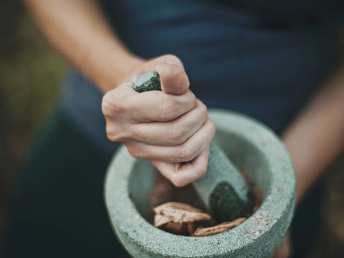 Hands hold a pestle, grinding herbs and possibly roots in a stone mortar, suggesting the preparation of traditional medicine or remedies.