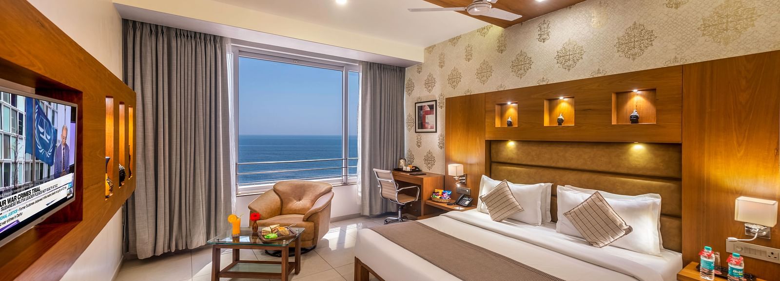 A  room featuring a comfortable bed, an armchair and coffee table by a window with an ocean view, a work desk, and a wall-mounted television.