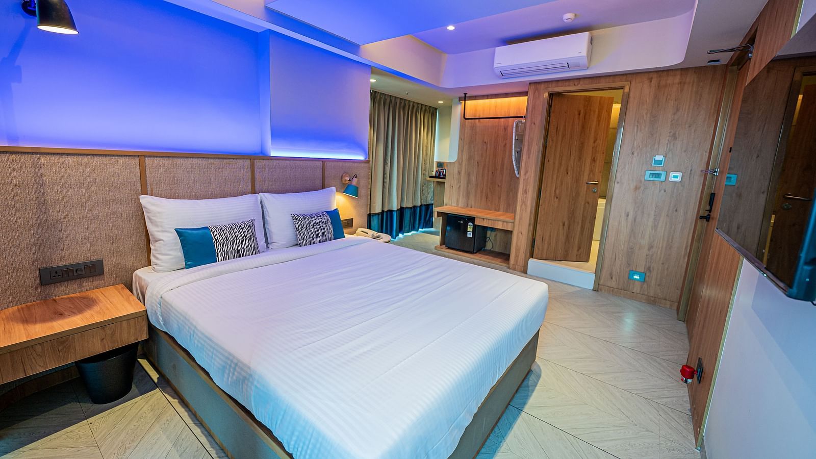 A spacious room at Indie Stays Mumbai International Airport features a large bed, a wooden bedside table, a wall-mounted TV, and a wooden door leading to another room.