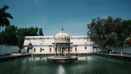 A view of a fountain inside a waterbody with a building in the background surrounded by trees at Saheliyon ki Bari.