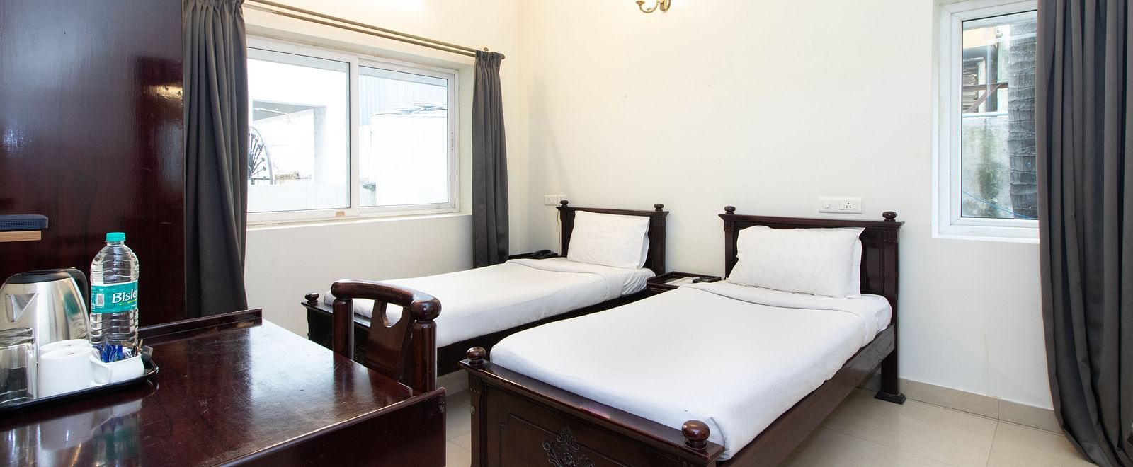 A room showcasing twin beds with white bedding, a work desk, a chair, a tea and coffee maker, a bedside table, and a window at Sanctum Suites Indiranagar, Bangalore.