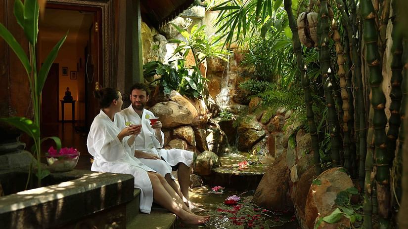 Two people in bathrobes enjoying beverages in martini glasses while dipping their feet in the shallow stream at Uday Samudra Leisure Beach Hotel