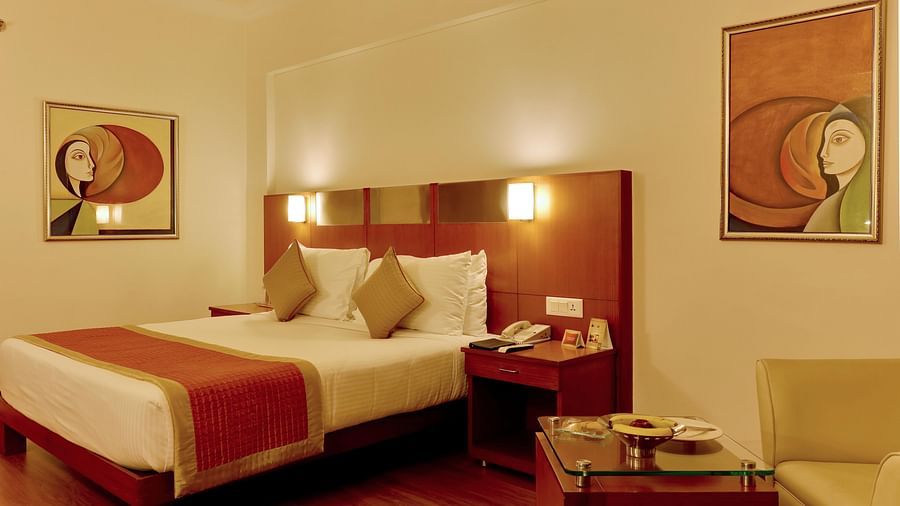 An image of a Deluxe Room with double size bed, bedside table and lam on either side and a huge window covered with curtains - The Suryaa New Delhi