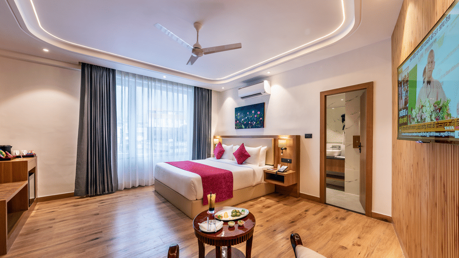 An expansive guest room at Lords Inn Katra featuring a king size bed, an armchair, a television, a wardrobe, and large draped windows.