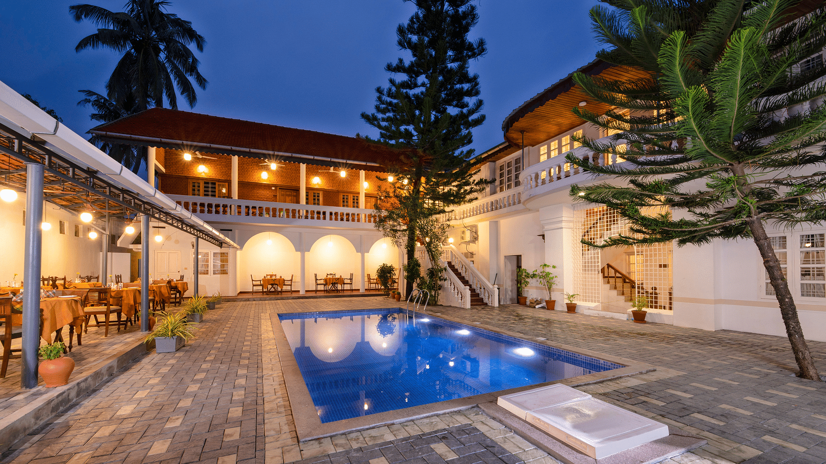 A courtyard with a swimming pool, surrounded by a colonial-style white building with arched windows, warm lighting, and lush greenery. - Abad Dutch Bungalow, Elphinstone Road, Fort Kochi