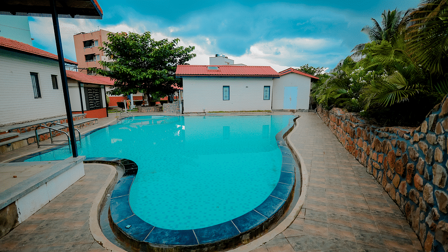 A straight angle view of a uniquely shaped swimming pool at Nirantara Resort & Spa, Bengaluru, surrounded by greenery and buildings.