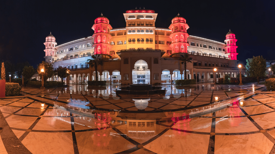 A view of the best hotel in Nathdwara, Shri Vallabh Vilas Lords Plaza, illuminated in pink lights at night, with symmetrical towers, a central fountain, and its reflection in the patterned floor in the foreground.