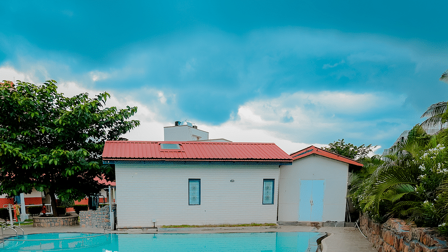 A straight angle view of a uniquely shaped swimming pool at Nirantara Resort & Spa, Bengaluru, surrounded by greenery and buildings.