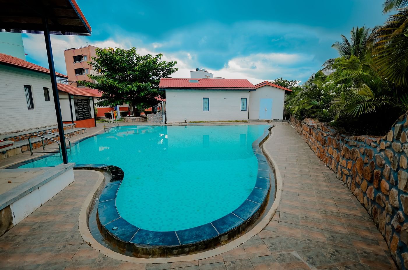 A straight angle view of a uniquely shaped swimming pool at Nirantara Resort & Spa, Bengaluru, surrounded by greenery and buildings.