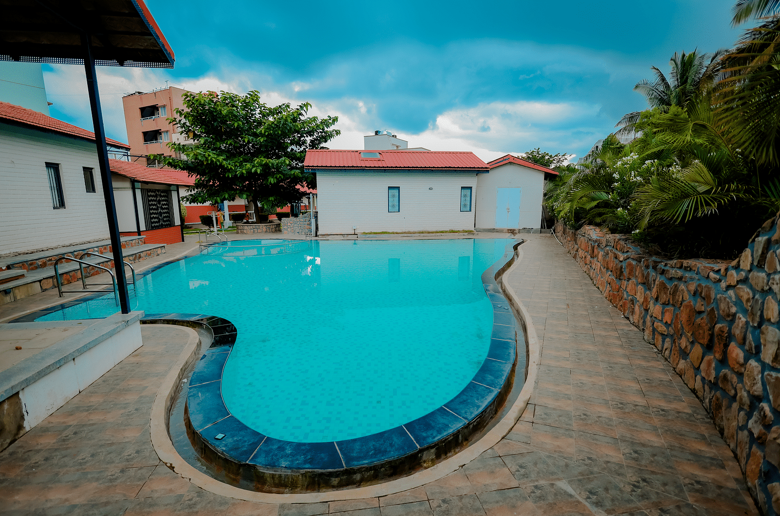 A straight angle view of a uniquely shaped swimming pool at Nirantara Resort & Spa, Bengaluru, surrounded by greenery and buildings.