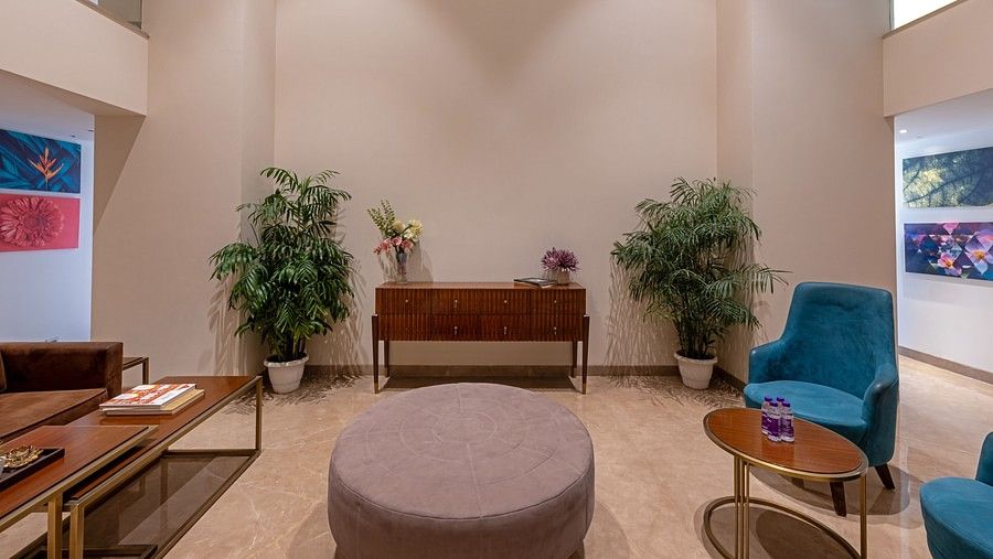 The lobby’s central seating arrangement at Hotel Saket 27 features a round table flanked by two coffee tables, with a brown table set against the wall. Small white pots with plants are thoughtfully placed in the corners, enhancing the room's aesthetic.