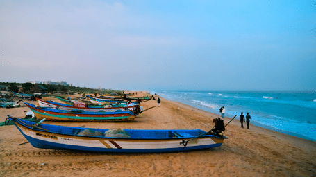 Colourful fishing boats lined along a sandy beach beside gentle waves, with visitors strolling the shoreline under a hazy sky