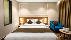 Deluxe Room at Pride Hotel Rishikesh featuring a large bed with a tan headboard with lamps on both sides over the nightstands and a sitting space arranged next to it by the window with a couch and a coffee table