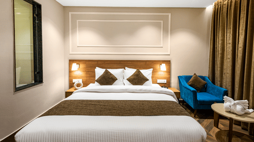 Deluxe Room at Pride Hotel Rishikesh featuring a large bed with a tan headboard with lamps on both sides over the nightstands and a sitting space arranged next to it by the window with a couch and a coffee table