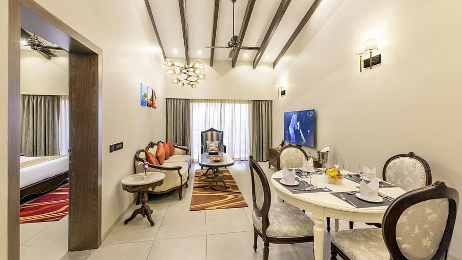 the wide angle shot of the dining area of our suite in candolim featuring a dining table with chairs, a couch with a coffee table, a flat screen television, and a door that opens to the bedroom - Golden Tulip, Candolim, Goa