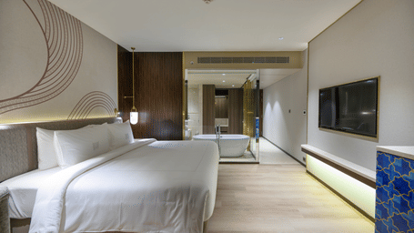 Bedroom with a plush bed with a patterned headboard facing the TV on the wall, and wooden flooring at Merlis Hotel, Coimbatore.