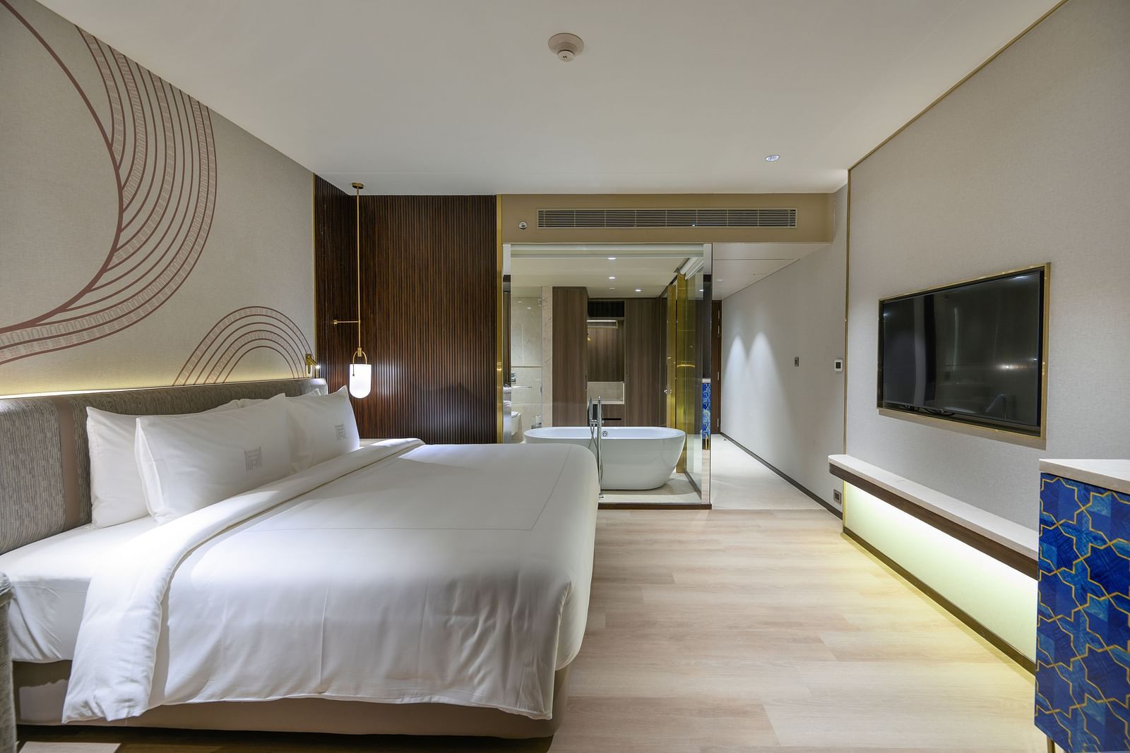 A bright hotel bedroom with a large bed, stylish headboard, and wooden flooring, suggesting a serene atmosphere at Merlis Hotel, Coimbatore.