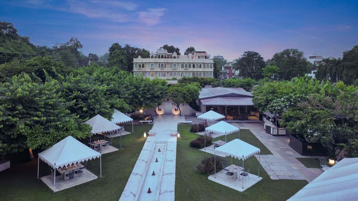 Exterior view of the back of Ram Pratap Palace in Udaipur, with a large garden, outdoor dining tents, and a central walkway at dusk.
