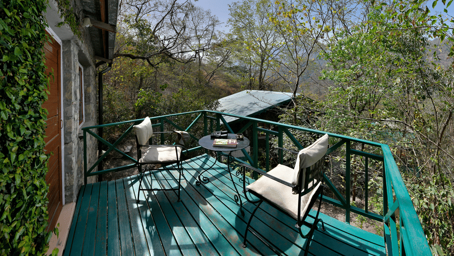 Private green wooden balcony with comfortable chairs and a small table surrounded by lush trees and forest foliage at Neemrana’s Glasshouse on The Ganges providing an intimate outdoor space for guests to relax and take in the fresh mountain air