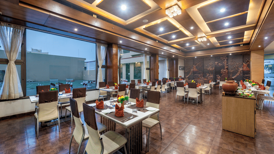 A spacious and elegantly designed restaurant with well-set tables adorned with flowers and large windows offering a view of the outdoors at Lords Inn Somnath.