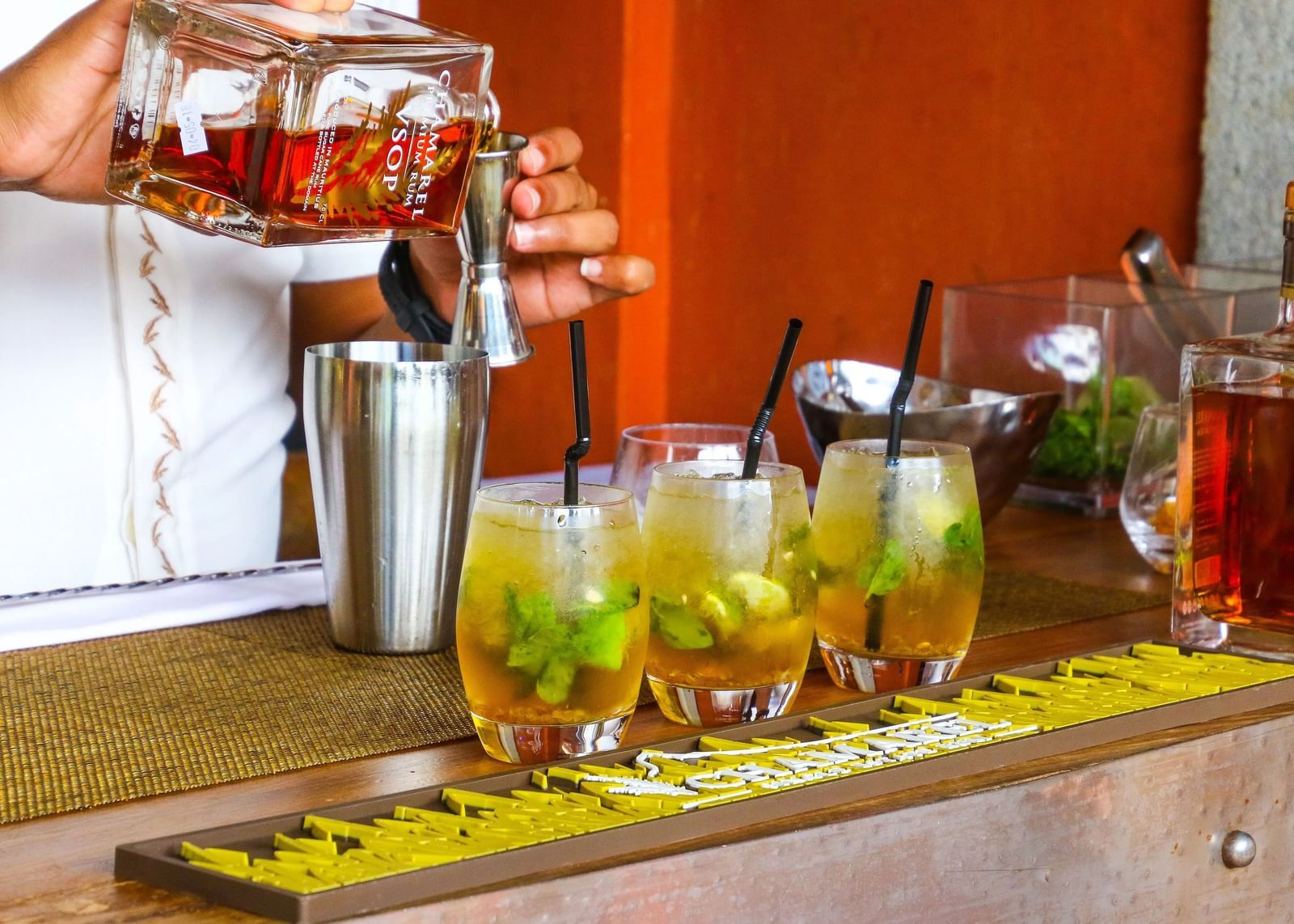 A bartender pours a drink into a glass filled with ice and garnishes, with other cocktails lined up on a bar.