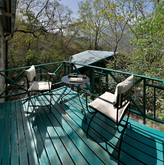 Private green wooden balcony with comfortable chairs and a small table surrounded by lush trees and forest foliage at Neemrana’s Glasshouse on The Ganges providing an intimate outdoor space for guests to relax and take in the fresh mountain air