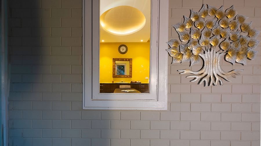 View of a decorative metallic tree wall art with golden leaves on a wall and a well lit room with yellow walls can be seen through the glass window.