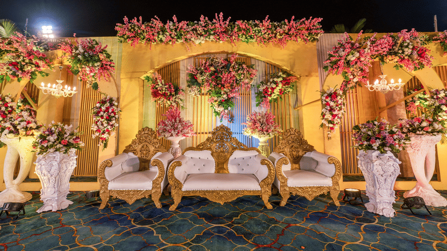 Multiple white and gold armchairs are placed on a stage decorated with yellow backdrops, floral arches, and white pedestals at Garh Govind.