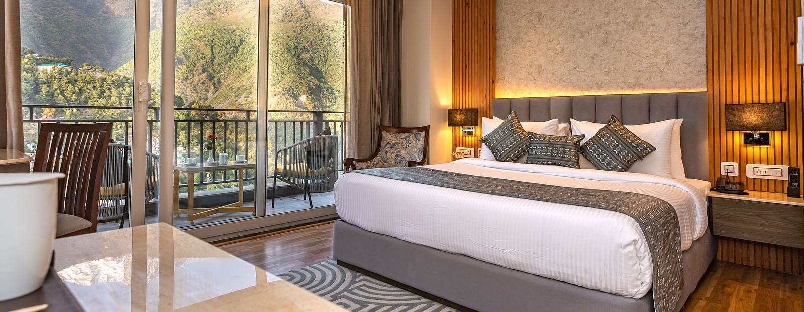 An image of a Suite with a bed, bed side table, lamp shades, and an expansive glass door leading to the balcony - The Belvedere Himalayan Retreat, McLeodganj