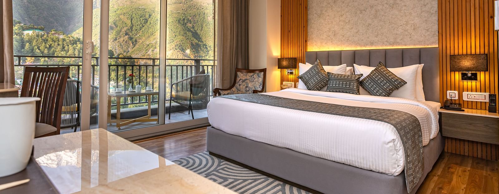 An image of a Suite with a bed, bed side table, lamp shades, and an expansive glass door leading to the balcony - The Belvedere Himalayan Retreat, McLeodganj