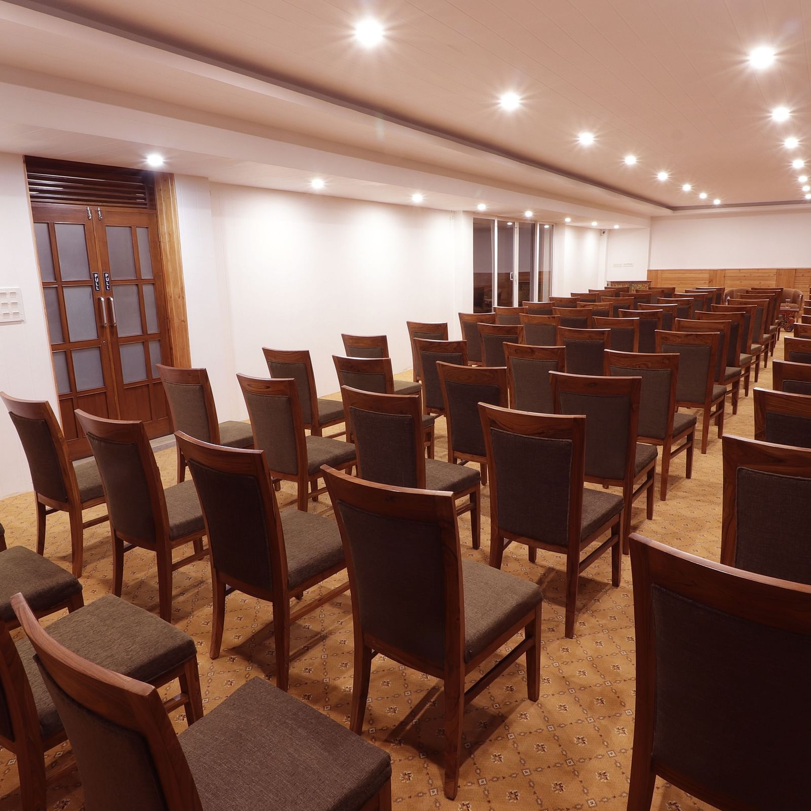 A narrow view of multiple wooden chairs arranged in rows under a well lit spacious hall at Denzong Lords Inn Gangtok.