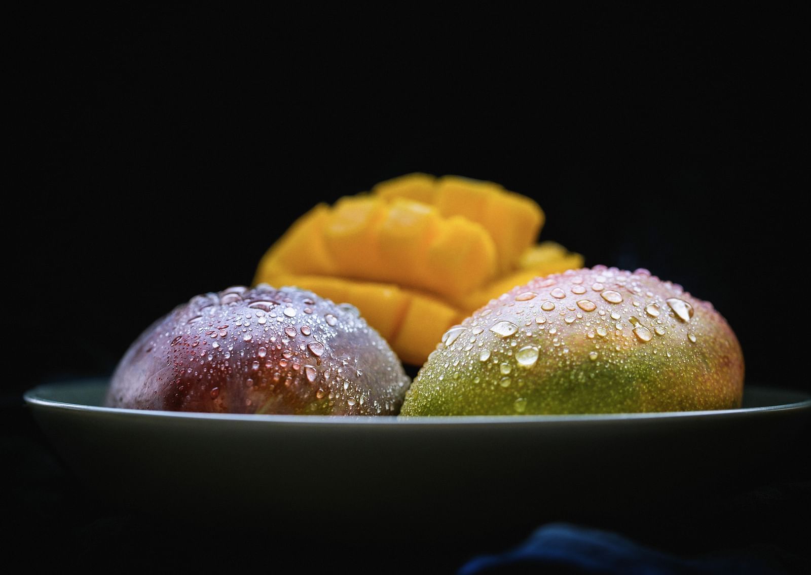 Two ripe mangoes and a cut mango kept in a bowl with water condensation on the mangoes.