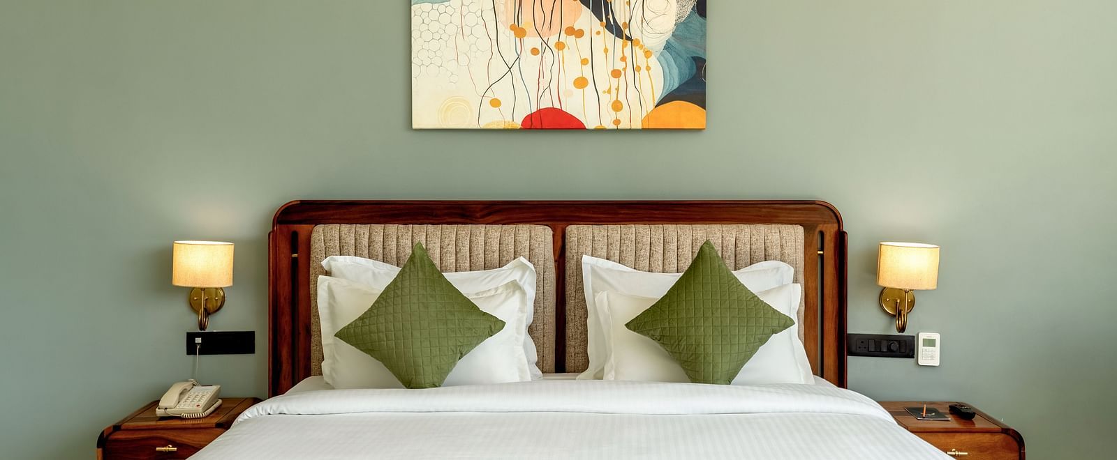 Pool View Room with a painting above a king size bed, bedside lamps, and green pillows at The Qualia Resort Club & Brewery, Udaipur.