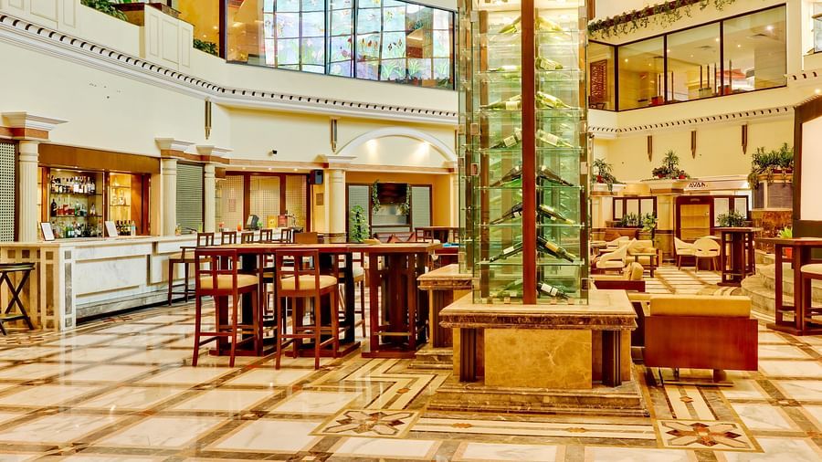 An image of bar with several sets of table and chairs and at the centre there is a multi storey glass cabinet where liquor bottles are kept - The Suryaa New Delhi