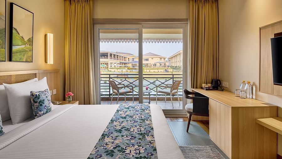A hotel room featuring a bed with a floral runner, a desk, a TV, and a view of a body of water through a sliding glass door.