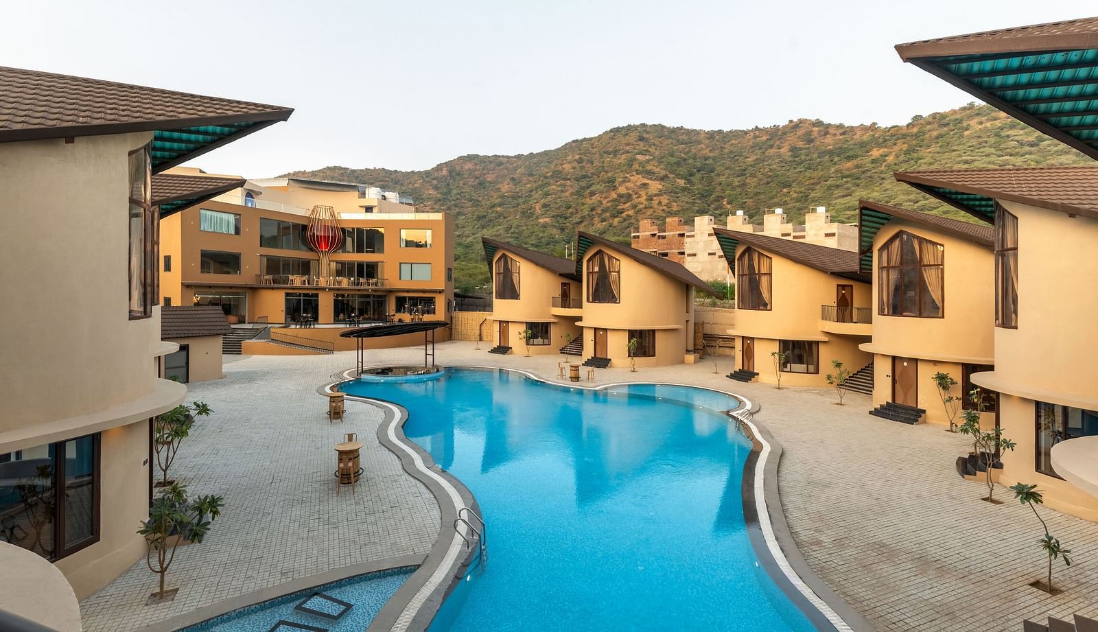 The Qualia Resort Club & Brewery, Udaipur with a blue swimming pool is nestled among buildings and hills, showcasing an outdoor leisure space.