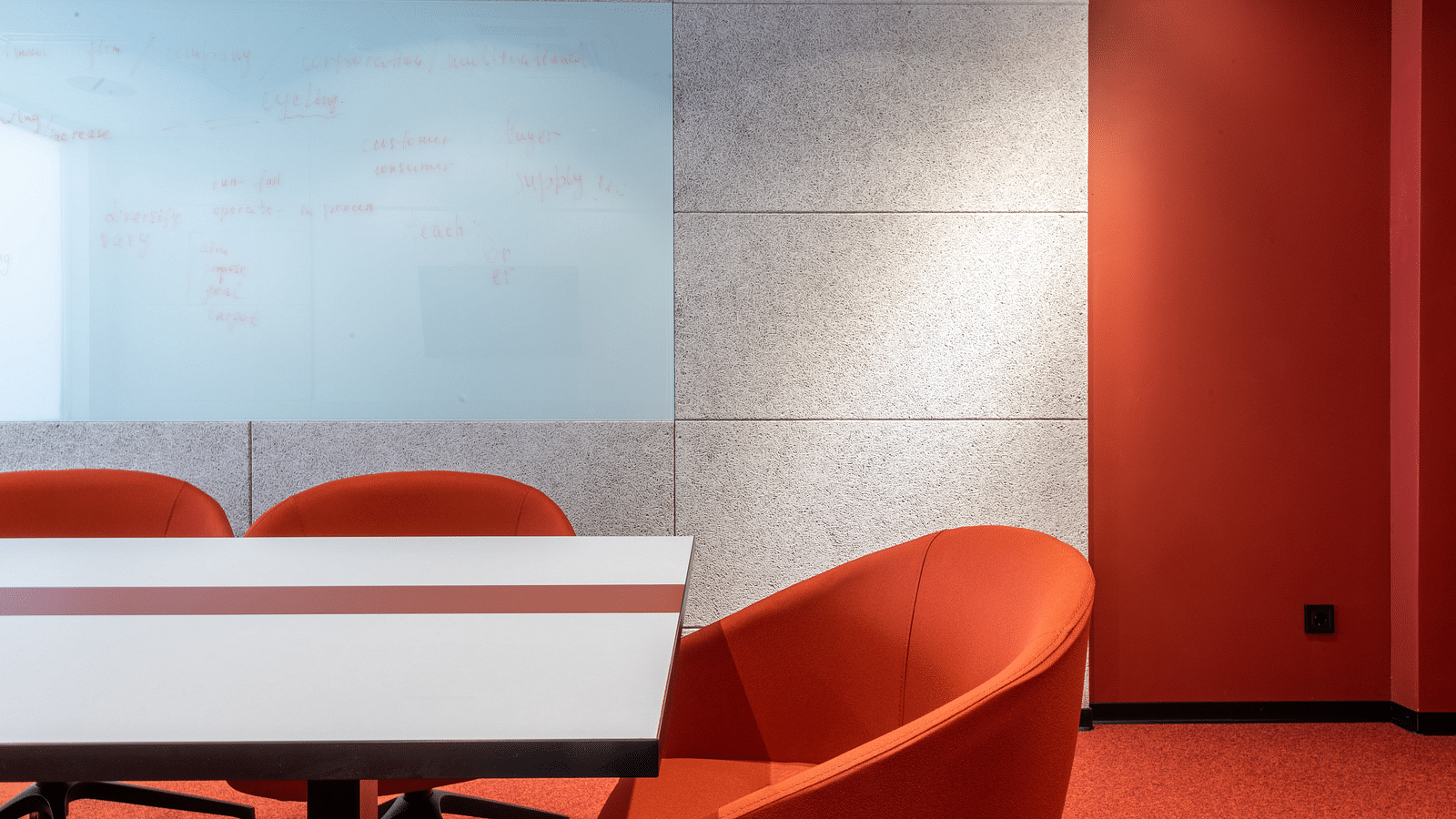 A close-up of a meeting room corner with a whiteboard filled with notes, a white and red table and red chairs on a red carpet