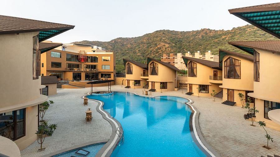 The Qualia Resort Club & Brewery, Udaipur with a blue swimming pool is nestled among buildings and hills, showcasing an outdoor leisure space.