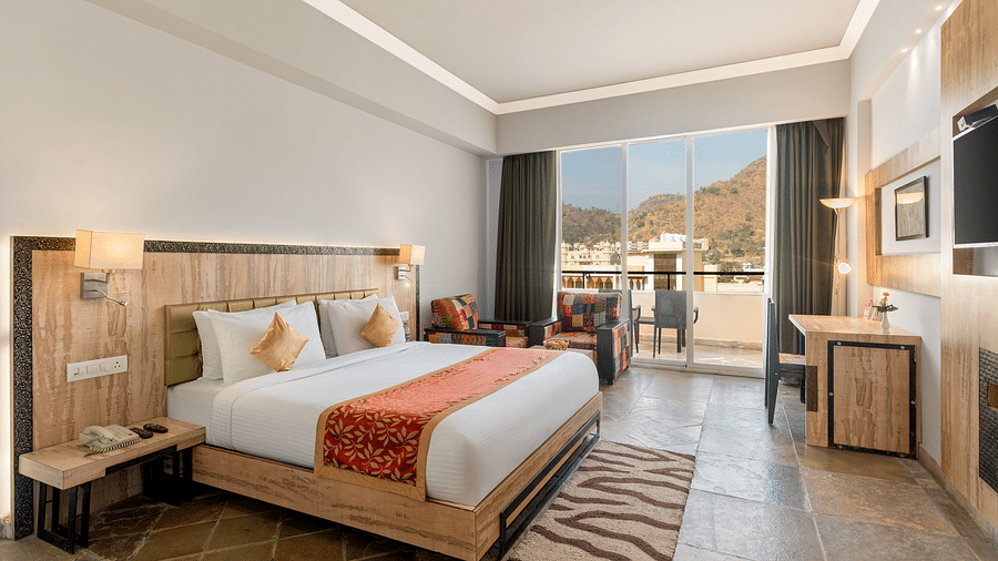 A hotel room with a made bed, desk, seating, and a balcony with a view - Via Lakhela Resort & Spa, Kumbhalgarh