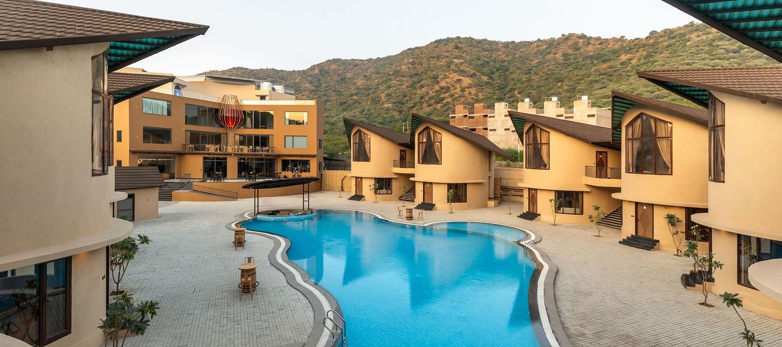 The Qualia Resort Club & Brewery, Udaipur with a blue swimming pool is nestled among buildings and hills, showcasing an outdoor leisure space.