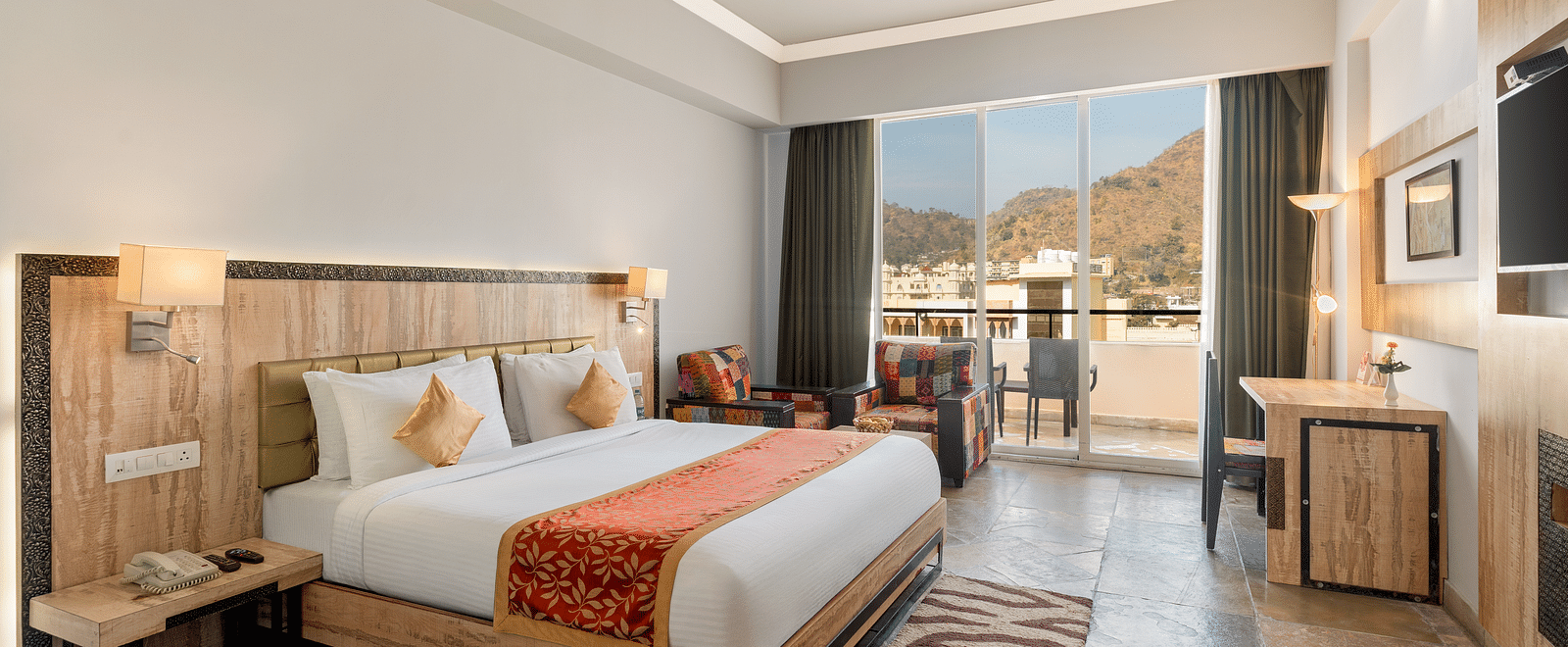 A hotel room with a made bed, desk, seating, and a balcony with a view - Via Lakhela Resort & Spa, Kumbhalgarh